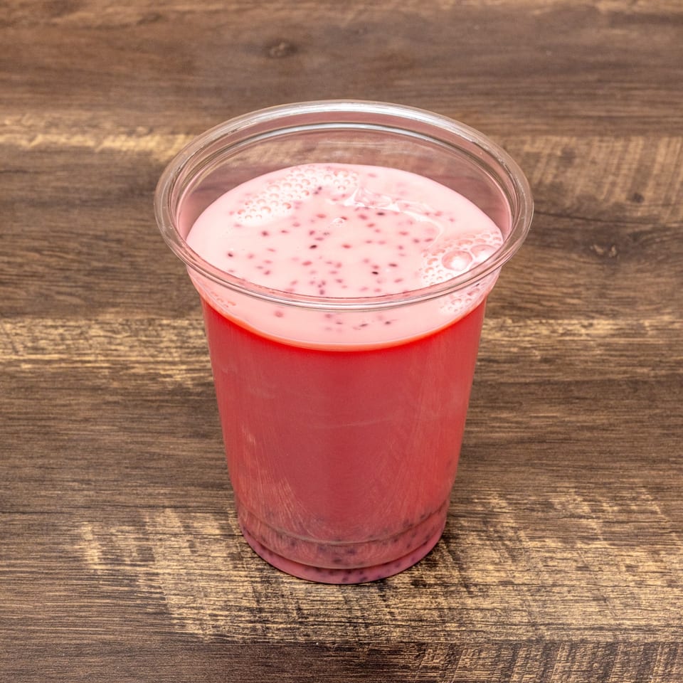 Pink falooda drink with black chia seeds and foam in a clear plastic cup on a wooden table.