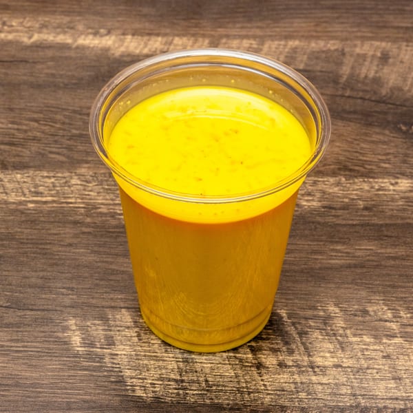 A vibrant yellow mango lassi in a clear plastic cup sits on a dark wooden surface.