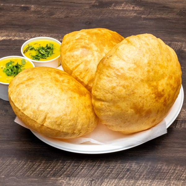 Three golden-brown Indian puri breads on a white plate with two cups of yellow curry.