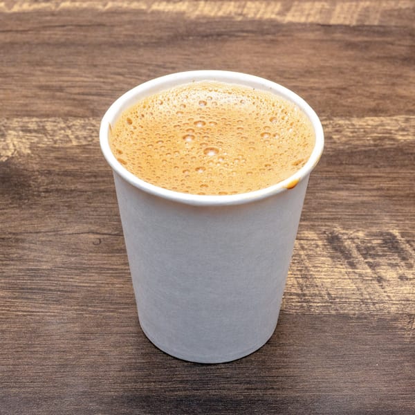A white paper cup filled with frothy, bubbly Indian filter coffee sits on a wooden table.